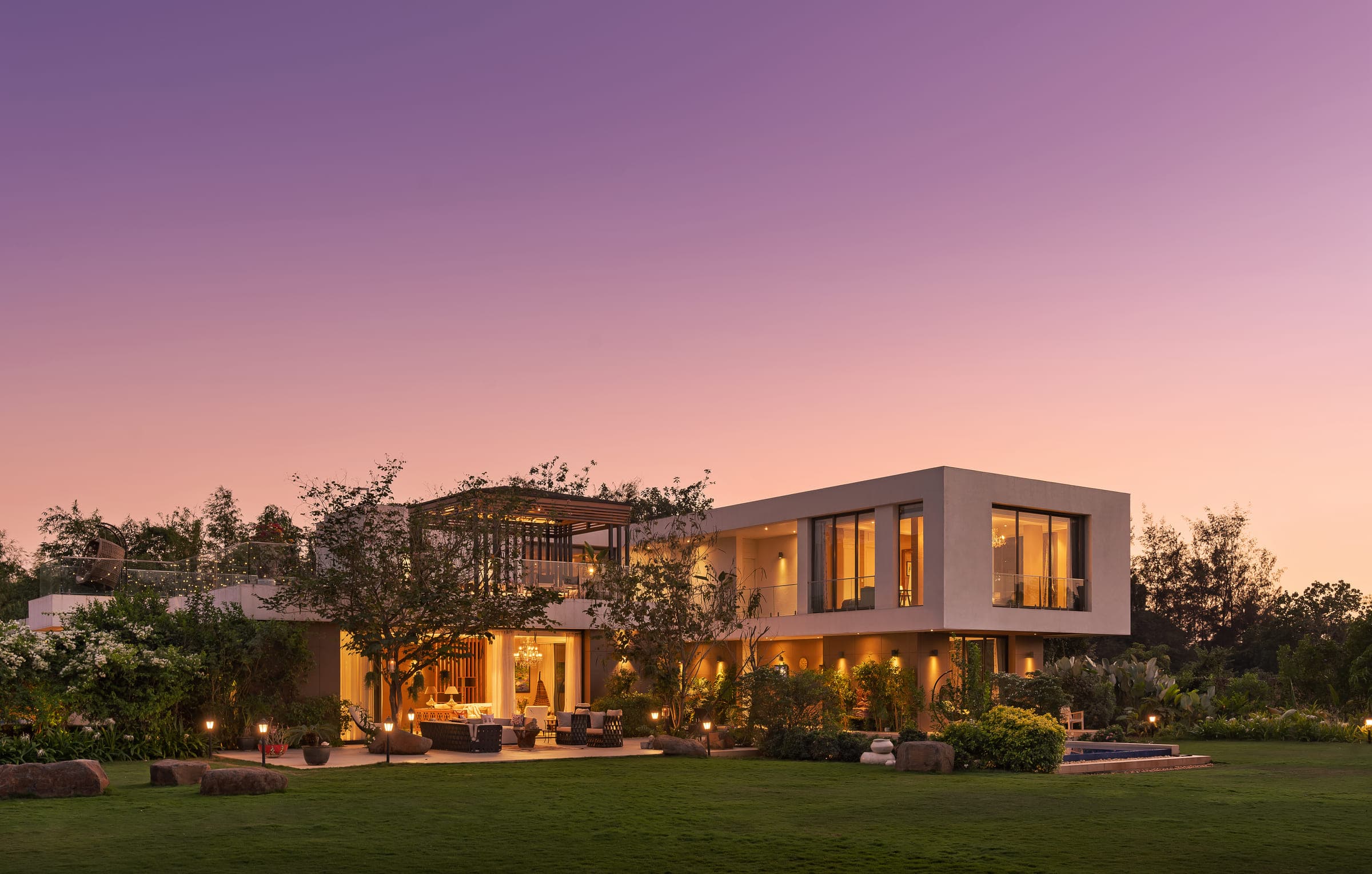 The house at dusk, seen across the lawn: low connected volumes with their windows lit warm against a violet sky, mature trees to either side.