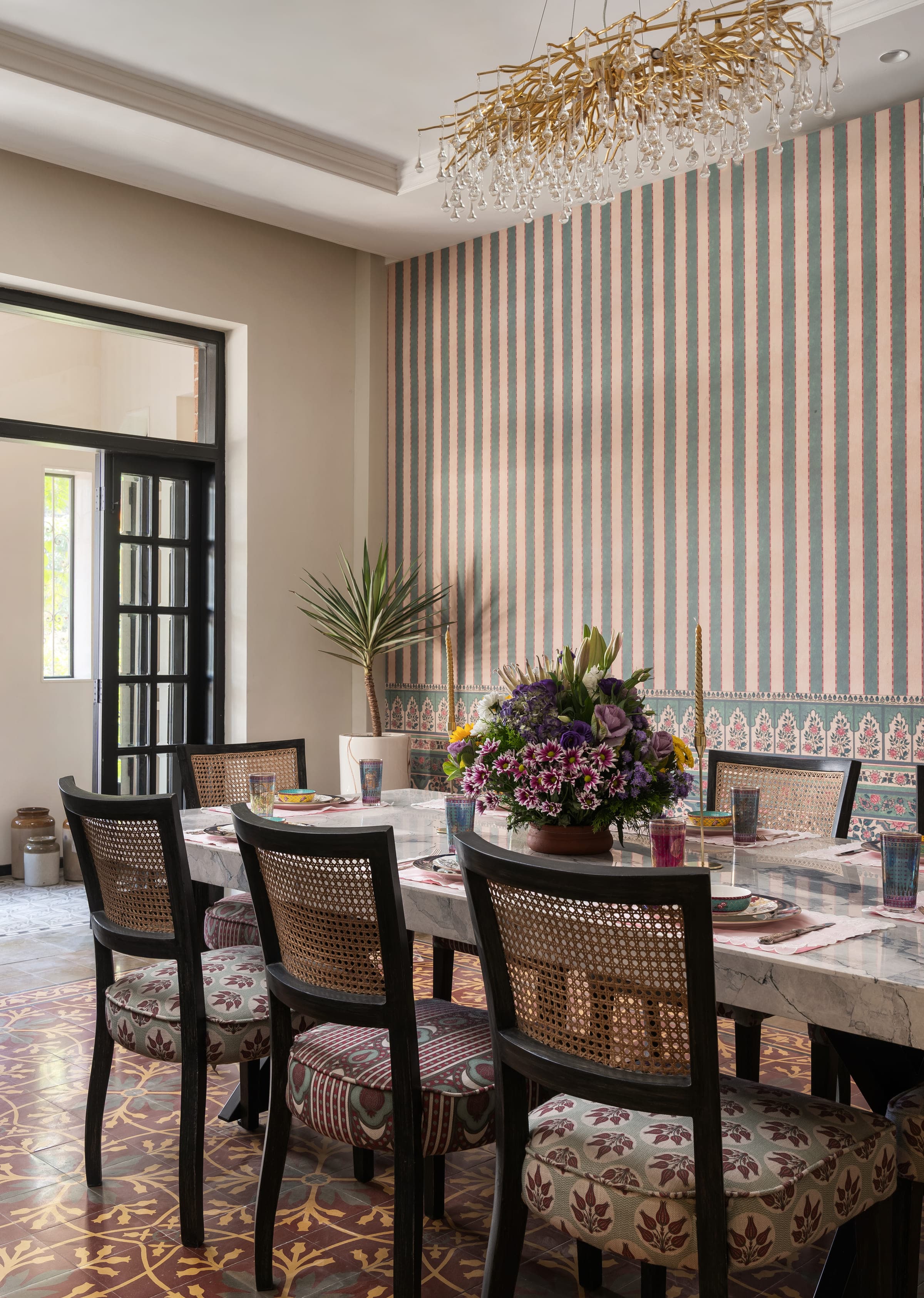 The dining room: a wall papered in sage and blush stripes above a floral border, a marble table laid for eight with cane-backed chairs on block-printed seats, a linear brass and crystal chandelier above, and the original patterned tile floor below.