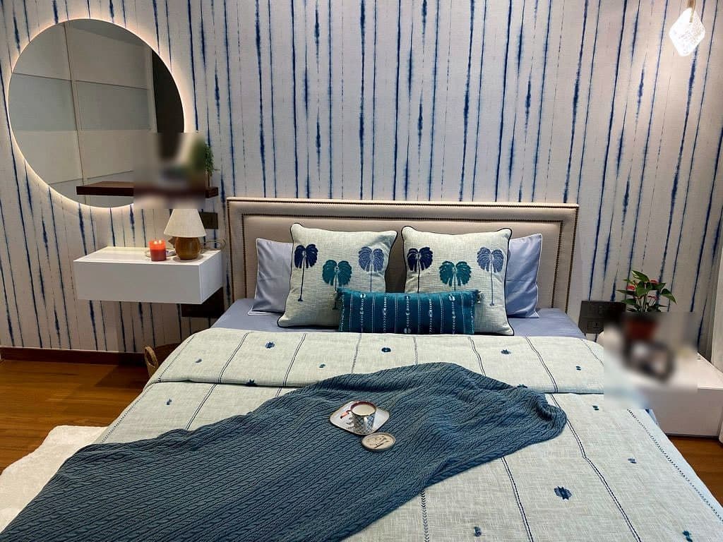 A bedroom papered in vertical indigo tie-dye stripes, a pale upholstered bed with printed cushions, and a round backlit mirror over a floating white console.
