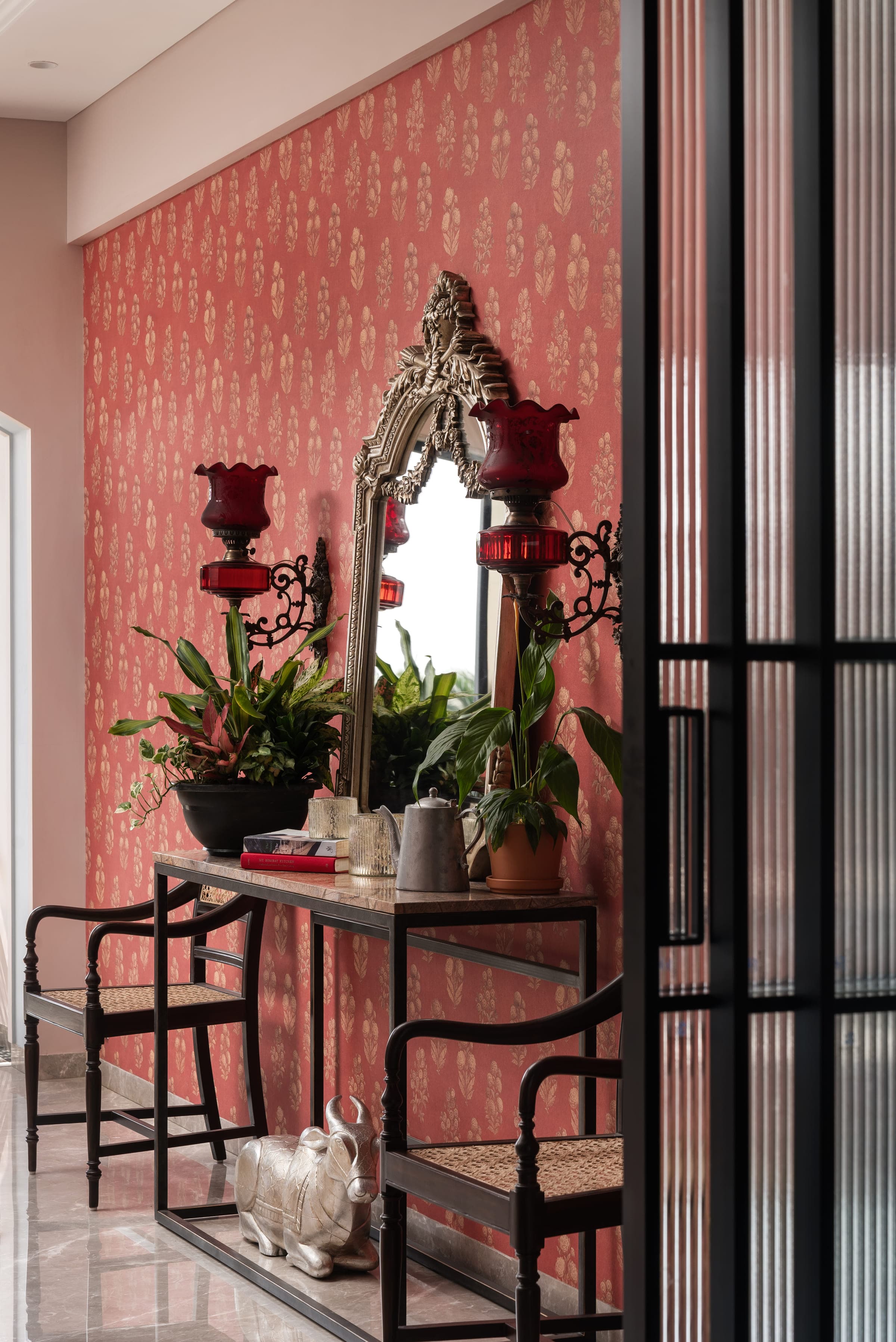 A wrought iron and marble console against a coral printed wall, with a gilt mirror above and ruby glass lamps at either end.