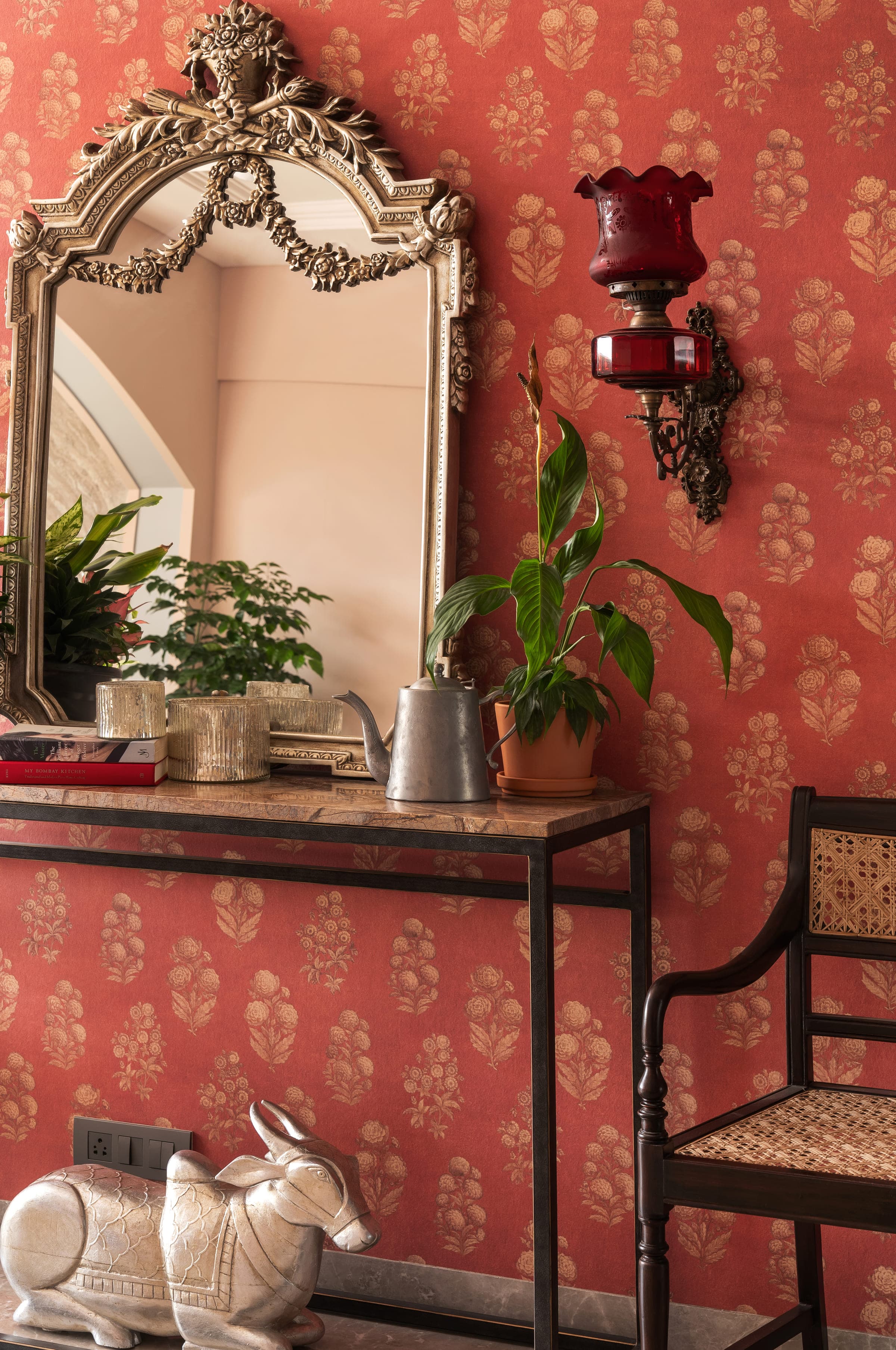 A close view of the wrought iron and marble console against the coral printed wall: a ruby glass lamp, a silver jug and a potted plant.
