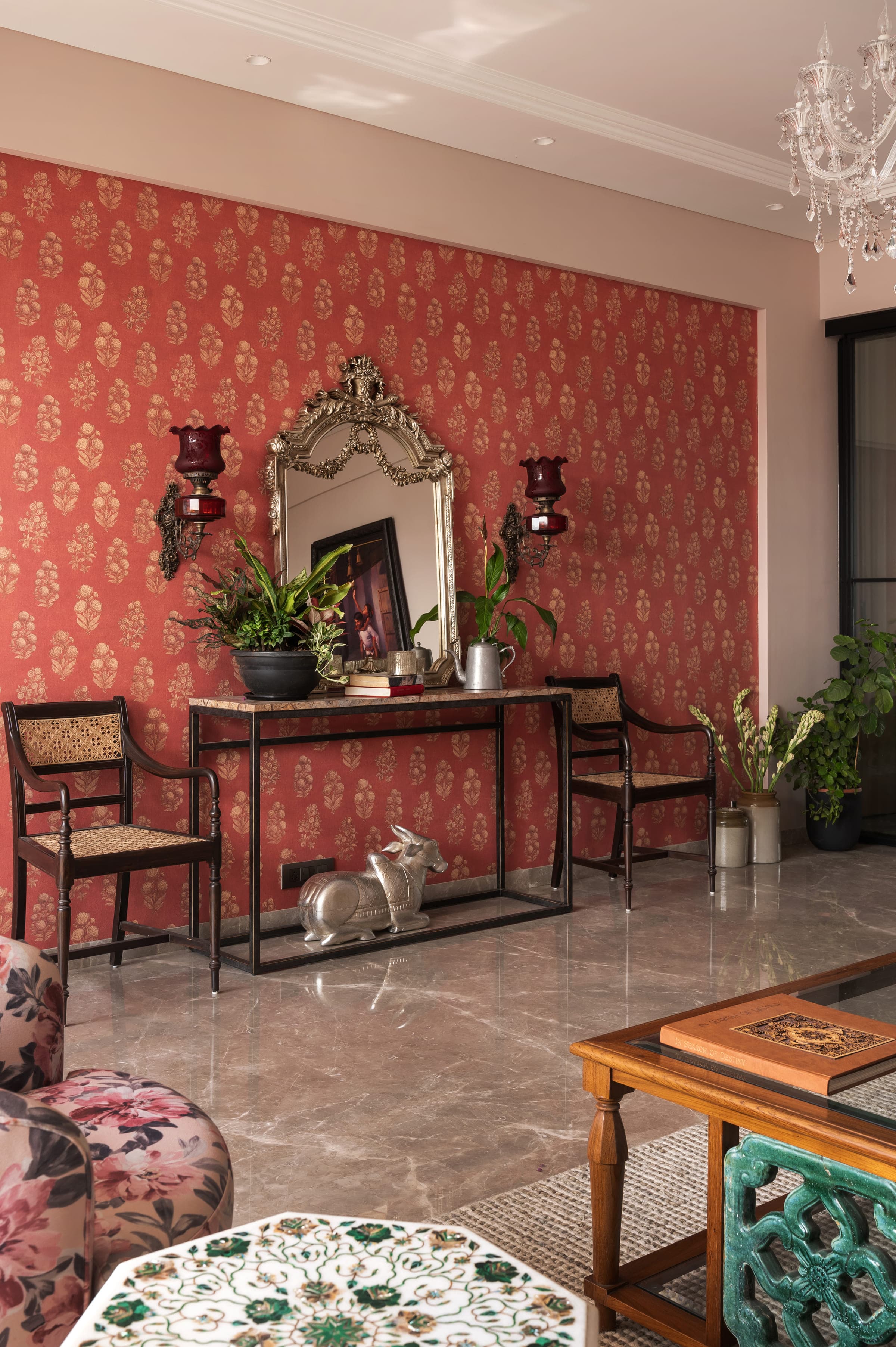 The coral printed wall seen wider, with cane-seated chairs to either side of the console and plants at floor level.