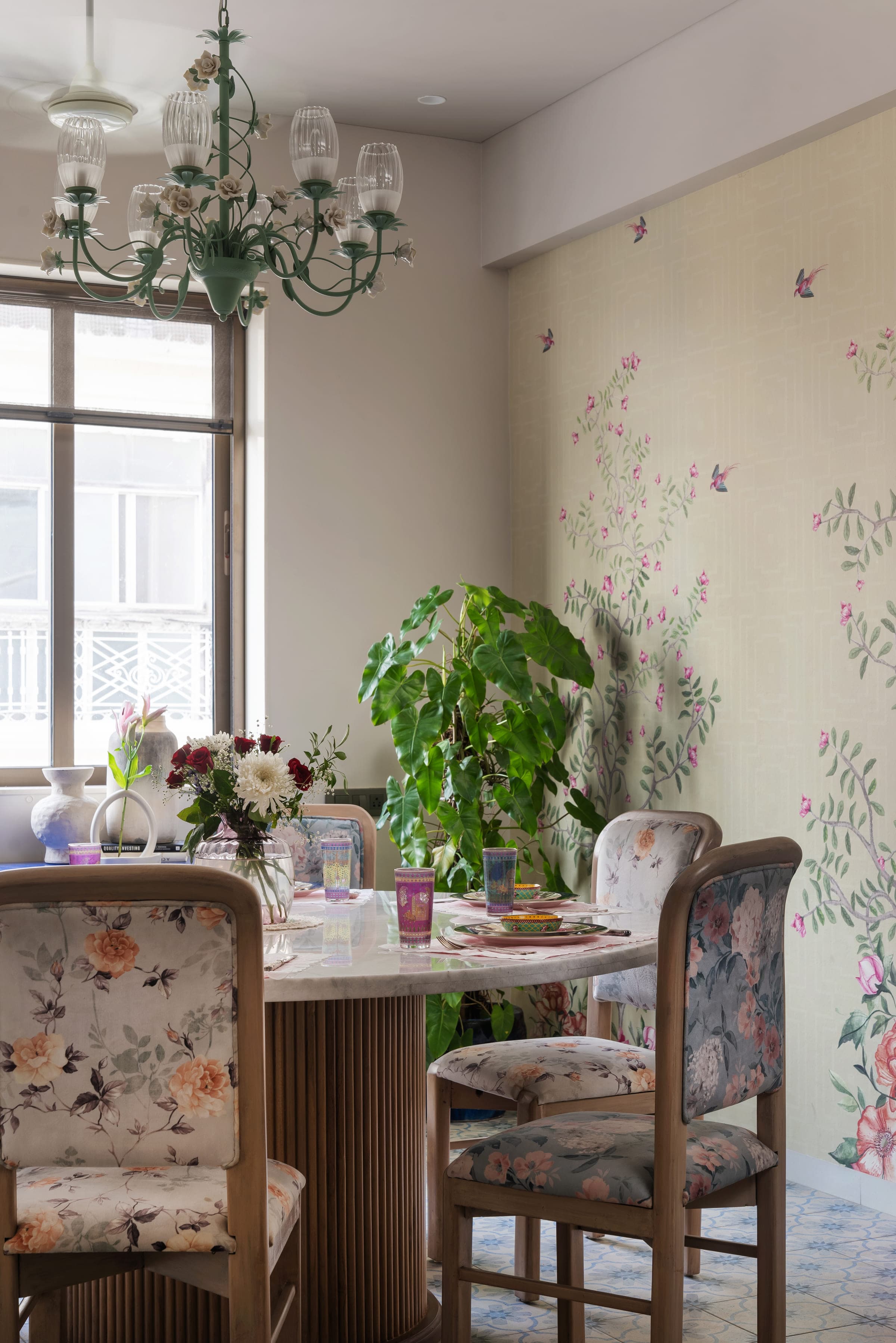 A dining table laid with coloured glassware in front of a wall printed with trailing foliage, plants on either side.