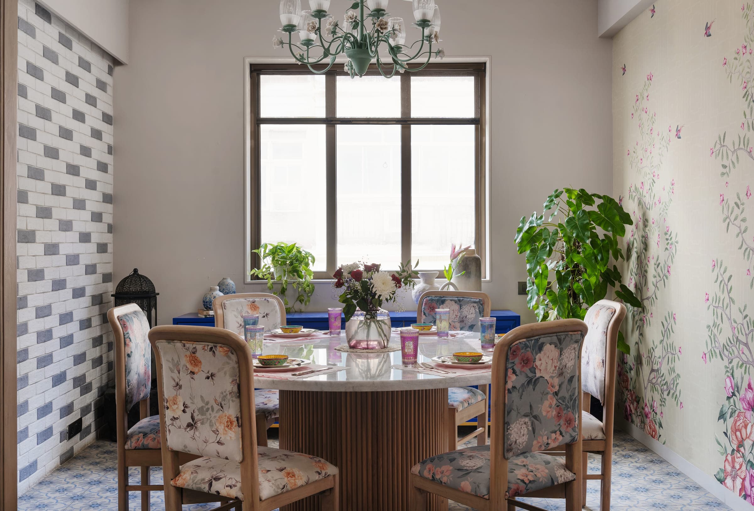 A round dining table and upholstered chairs beneath a chandelier, set in front of a tall window with a tiled screen wall alongside.