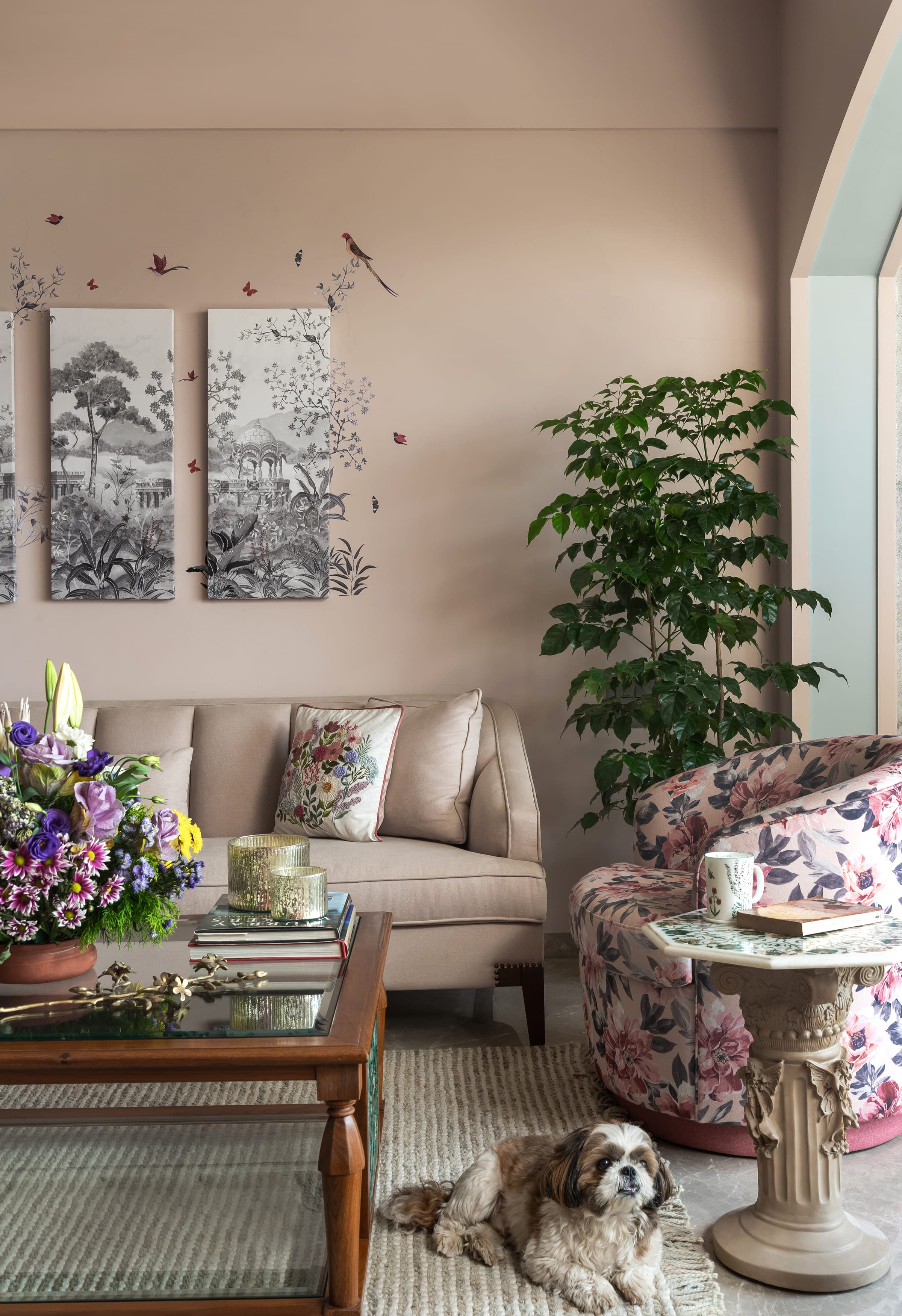 Cut flowers and a cake stand on a low table between pale sofas, a tall plant behind and framed prints on the wall.