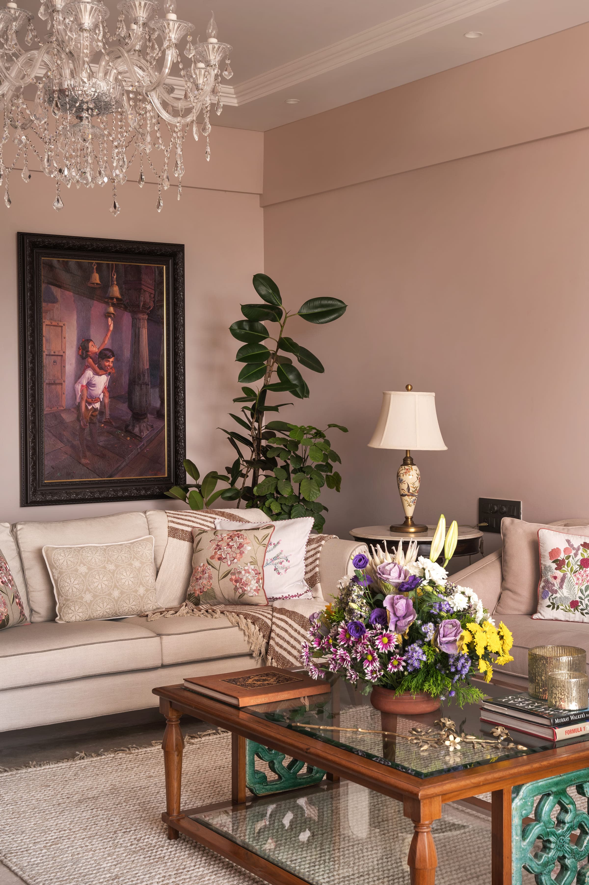 A living room corner with a large framed painting above a long sofa, a table lamp beside it and a tall fig in the corner.