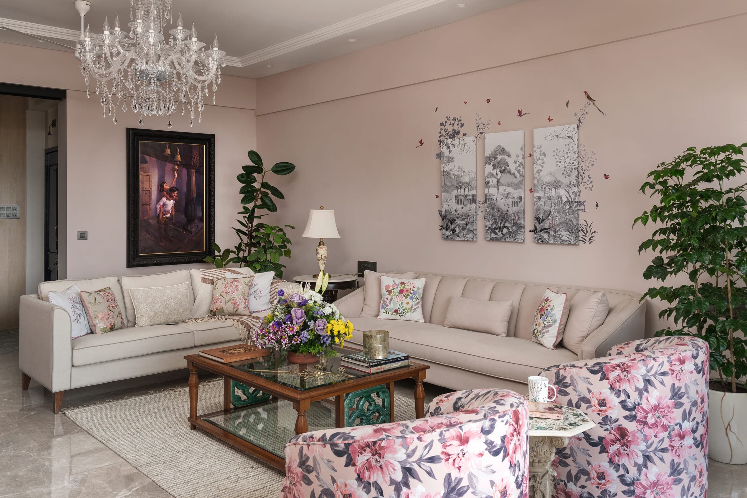 A pale living room with two long sofas facing each other across a glass table, floral armchairs at either end and a chandelier overhead.