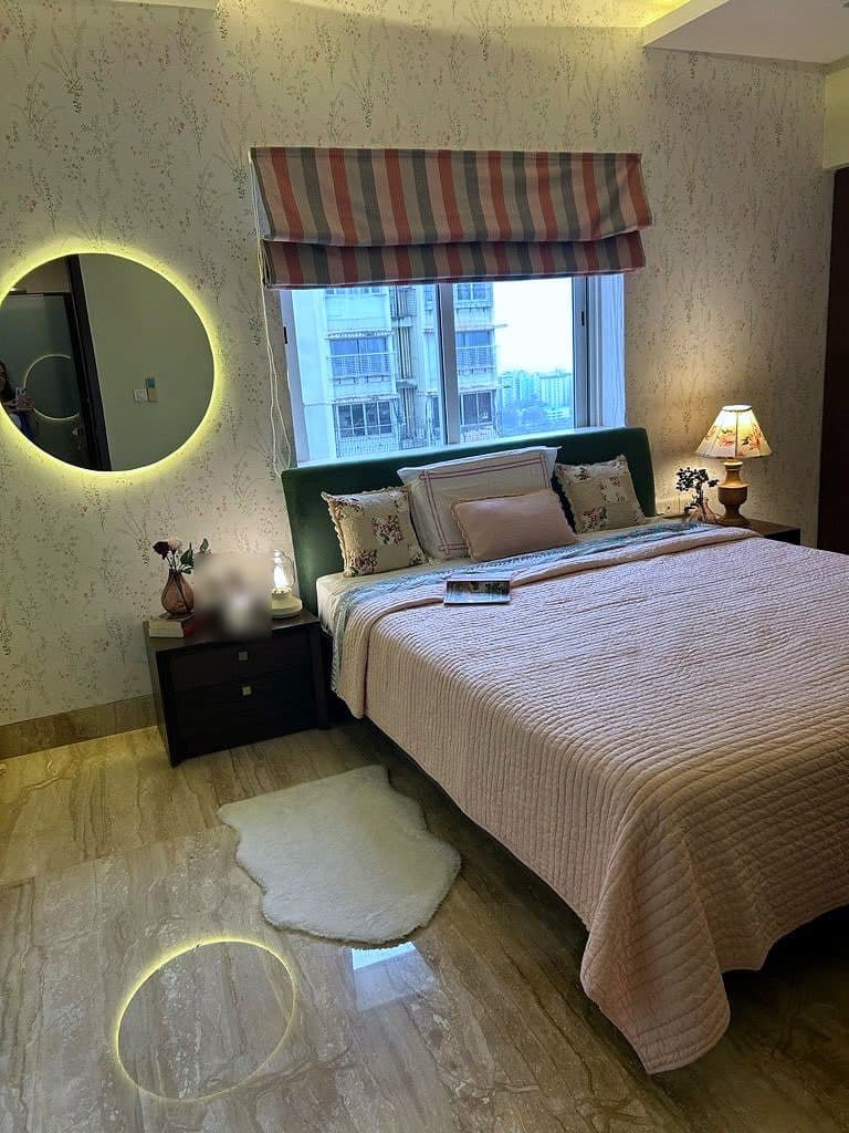 A bedroom with a deep green upholstered headboard, a round backlit mirror on the left and a striped blind over the window.