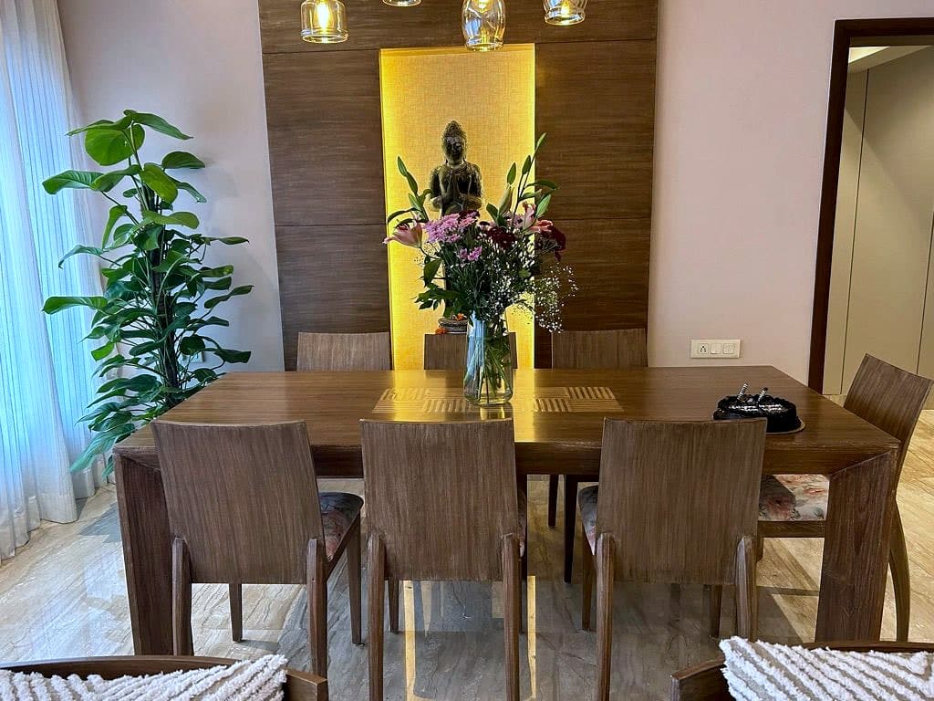 A teak dining table and six chairs facing a grained timber wall with a backlit recess holding a bronze figure, glass pendants above.
