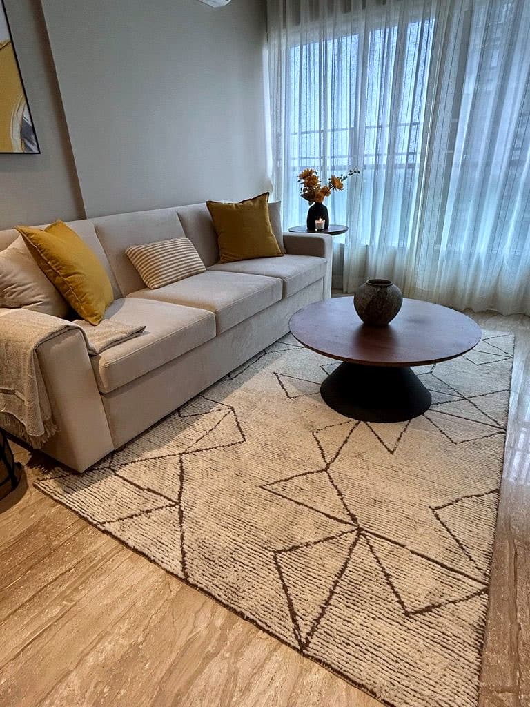 A long cream sofa with mustard cushions on a geometric flat-woven rug, a round timber table in front and sheer curtains behind.