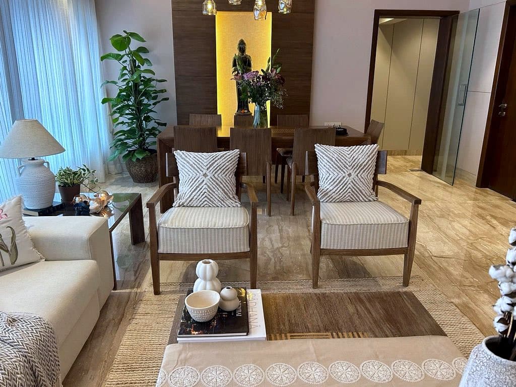 A living room opening into a dining room, cream armchairs in the foreground and a teak table beyond, set against a backlit alcove holding a bronze figure.