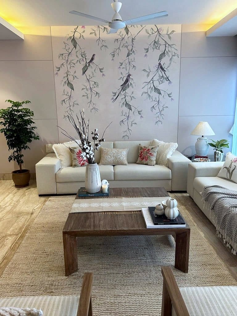 A living room with a hand-drawn wall panel of branches and birds behind two cream sofas, a low teak table and a potted ficus.