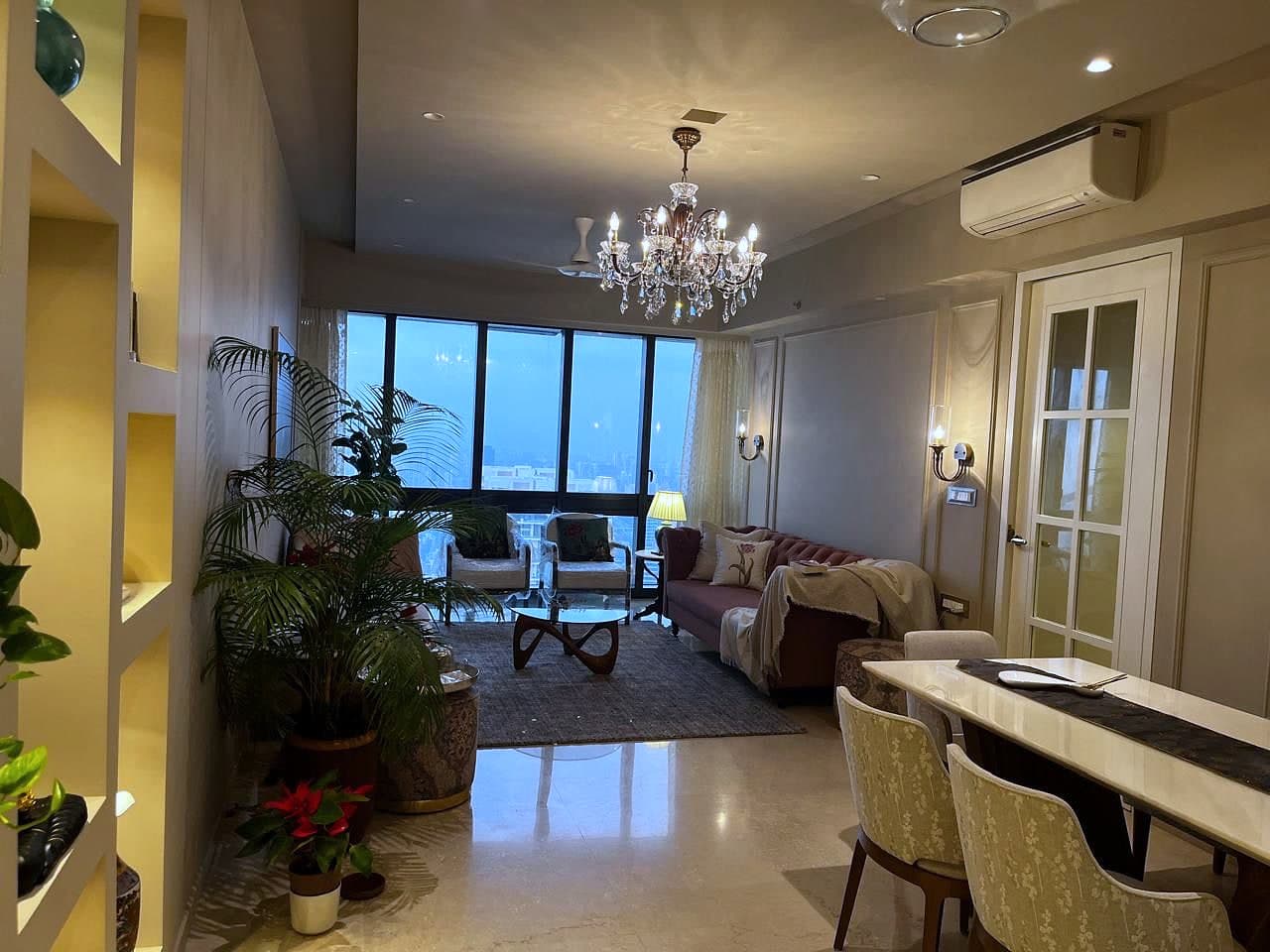 The apartment seen from the entrance at dusk: the dining table to the right, seating and the window wall beyond, a crystal chandelier over each end and planting at every corner.
