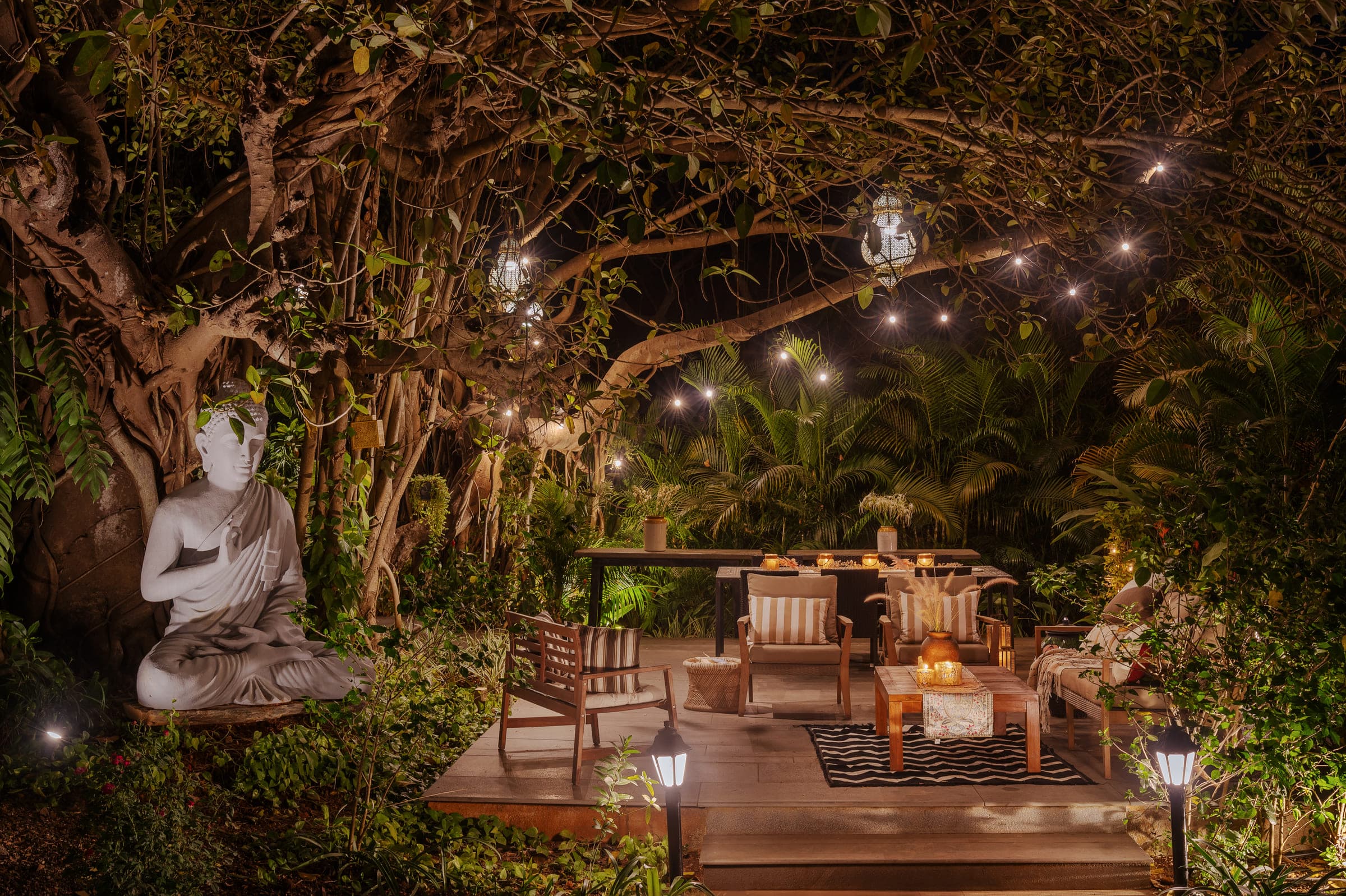 The deck at Private Villa in Alibaug after dark, strung with lights beneath the banyan, a stone Buddha at one end and timber seating set out under the branches.