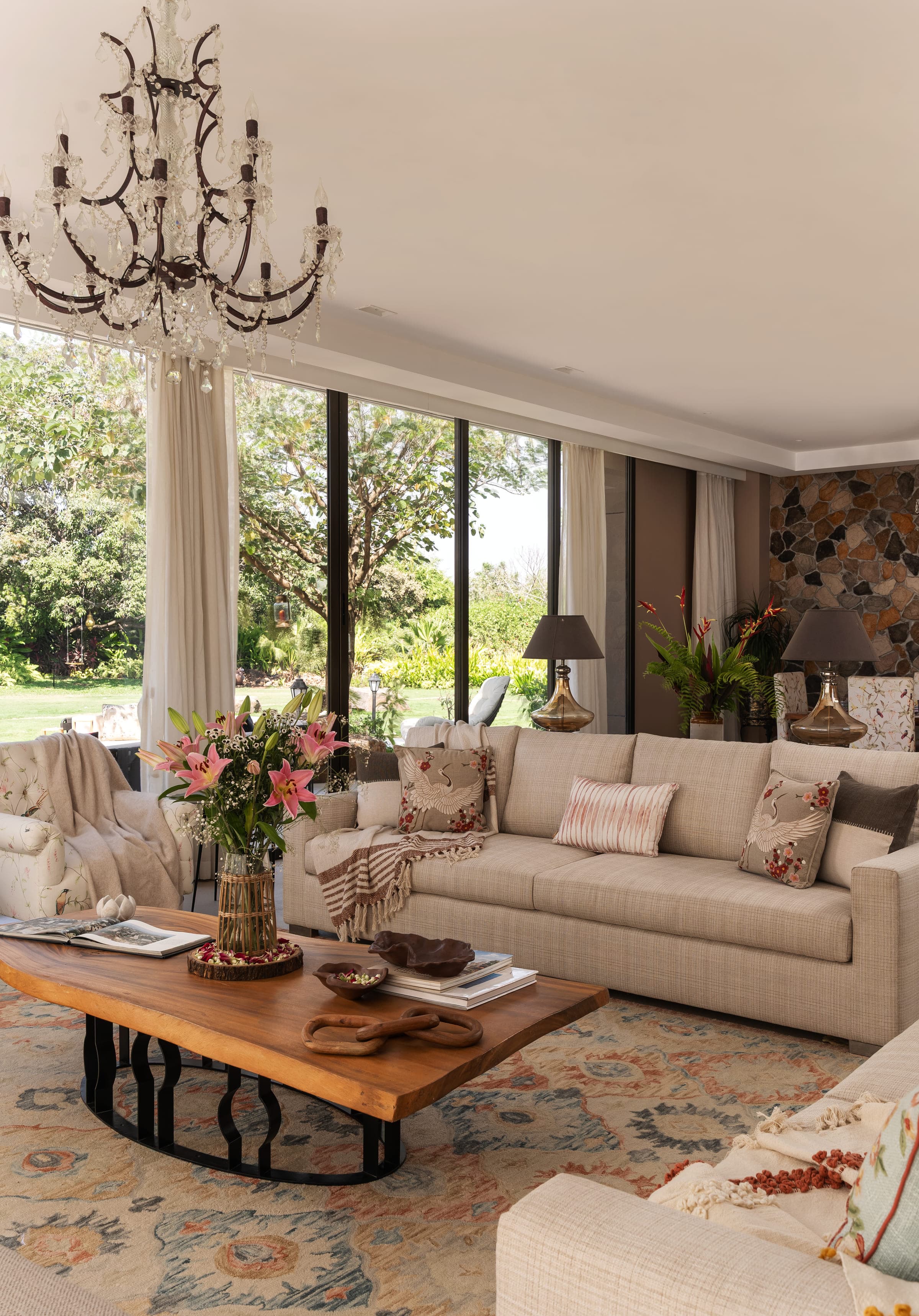 The living room: linen seating around a solid timber table, a chandelier overhead, a textured stone wall to one side and full-height glazing onto the garden.