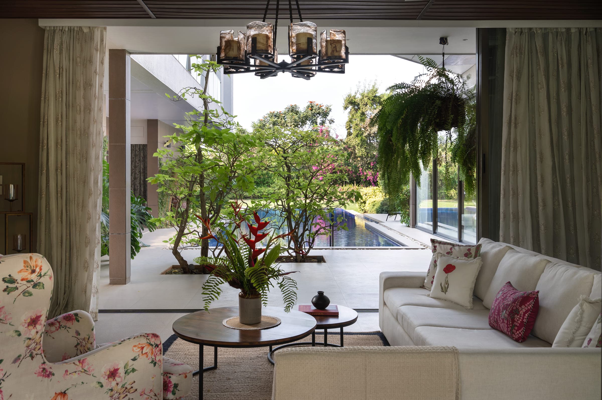 The living pavilion with its full-height glazing slid open onto the pool terrace, linen seating and a solid-log table inside, planting immediately beyond.