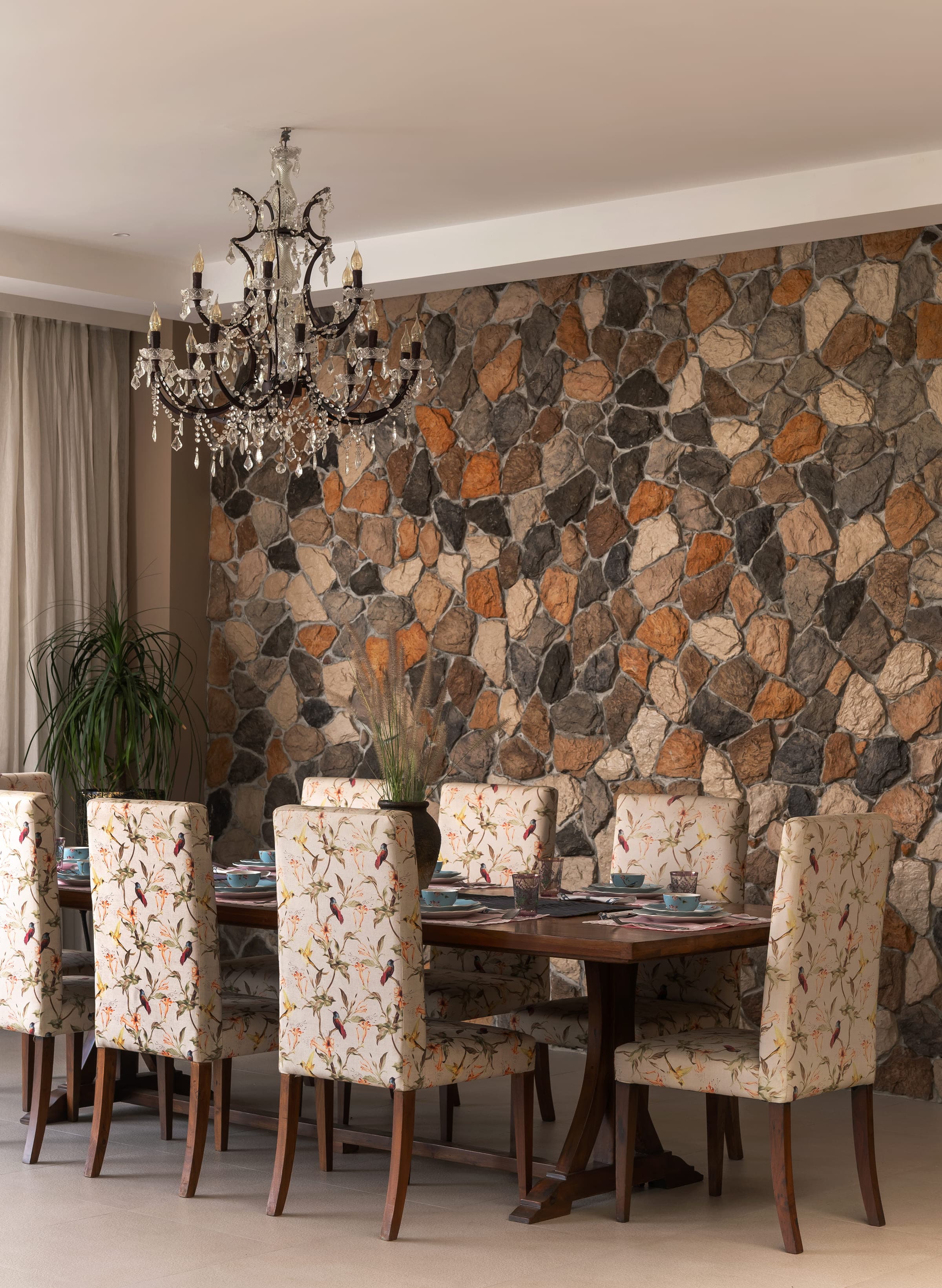 The dining table set against a wall of rough-cut stone stacked by hand in ochre, grey and cream, with a crystal chandelier above.