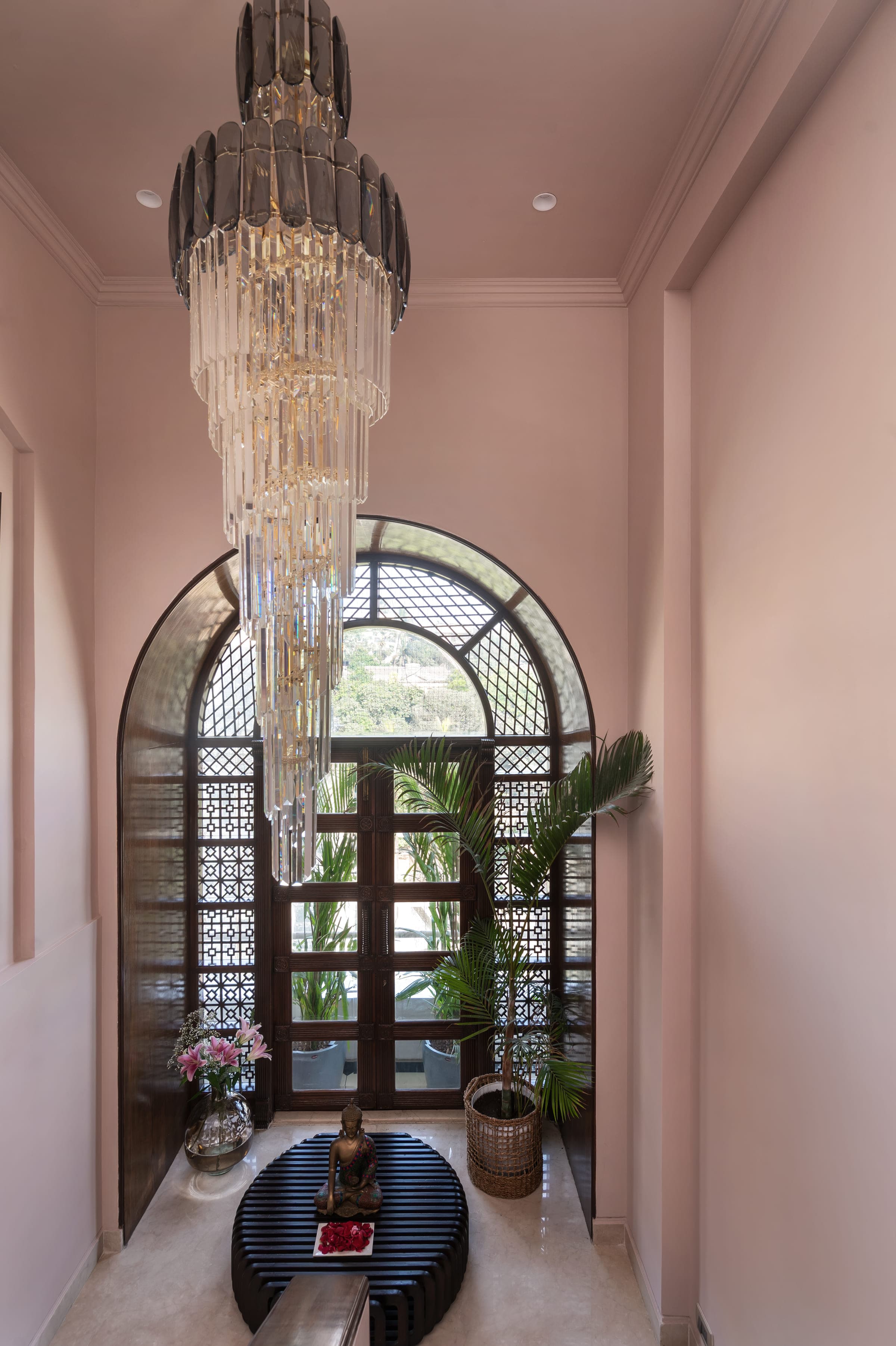 An arched window with a dark timber jali screen, a crystal chandelier hanging beside it against a blush wall.
