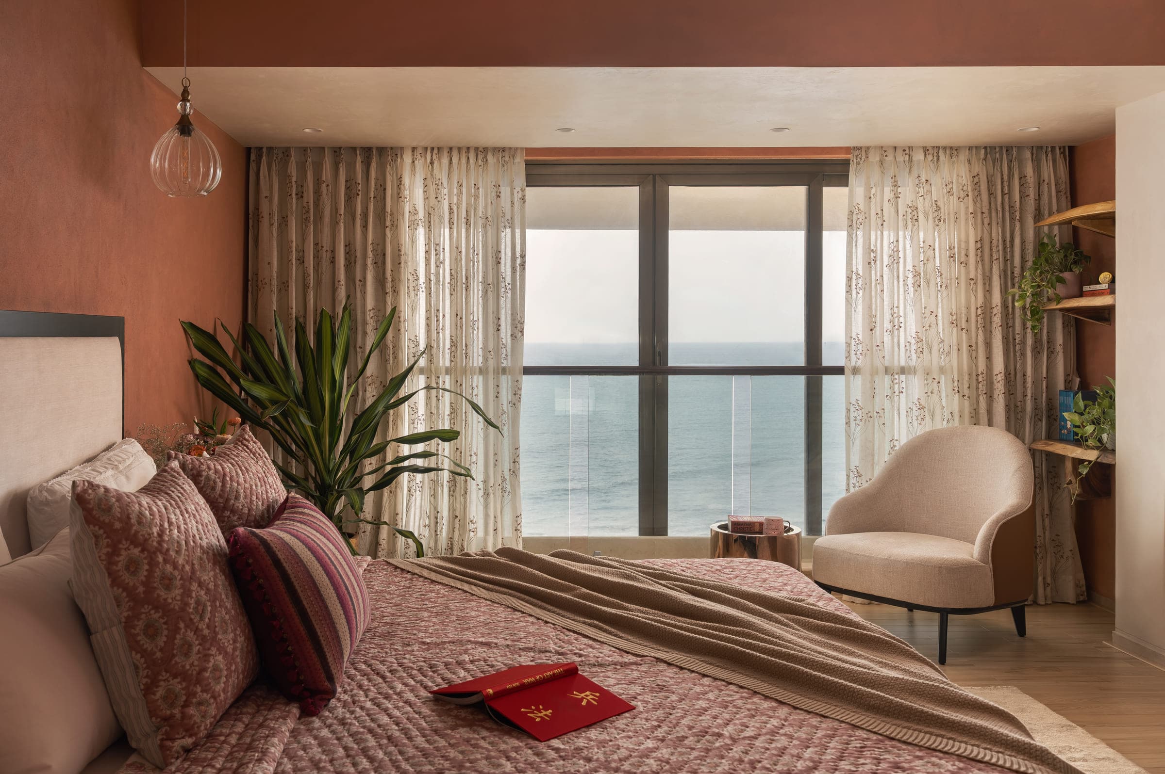 The bedroom after the work: terracotta walls, block-printed bedding, sheer curtains drawn back from a window filled with open water, and a low armchair beside it.