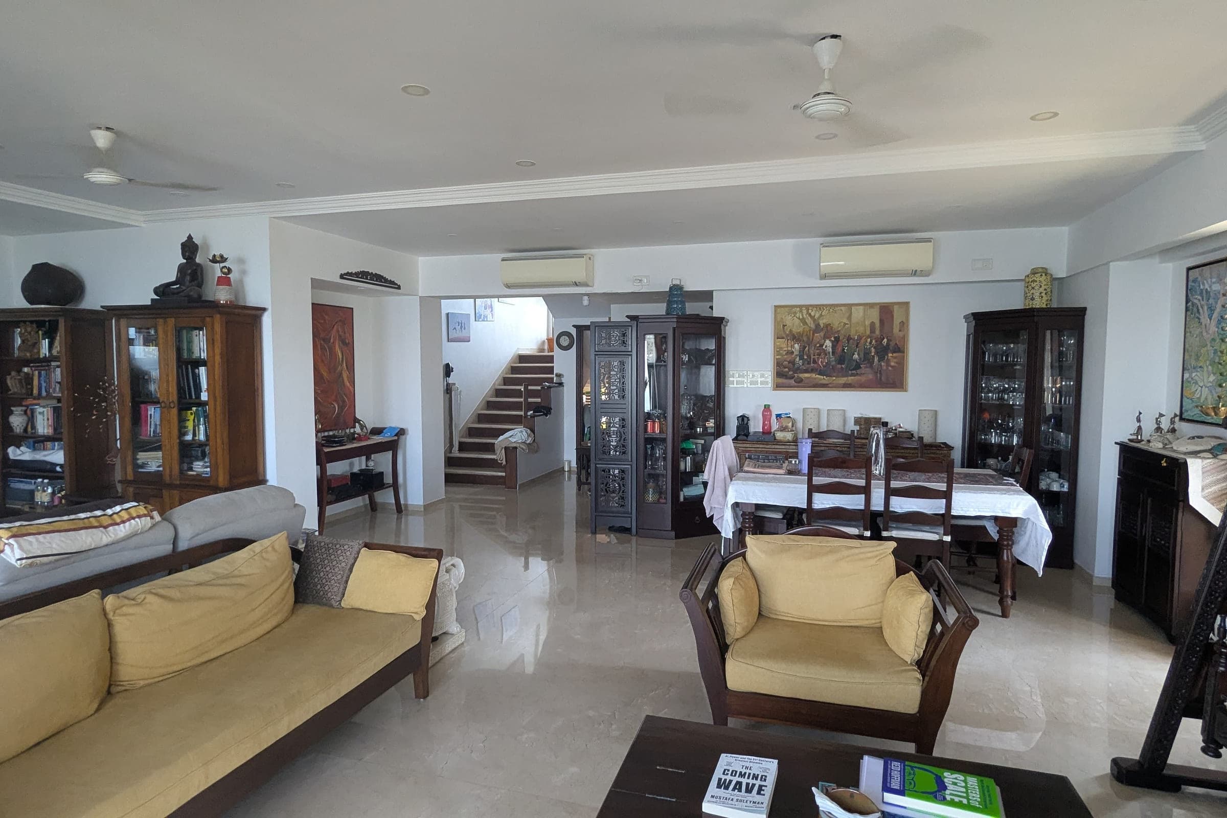 The living room before the work: carved dark wood cabinets and a display case against pale walls, yellow-cushioned settees, a marble floor and a staircase beyond.