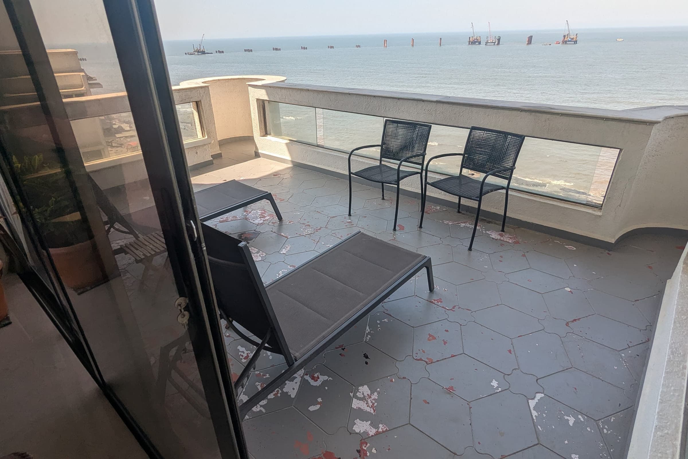 The terrace before the work: bare grey hexagonal tiles, two mesh chairs and a sun lounger pushed to one side, the sea beyond the parapet.