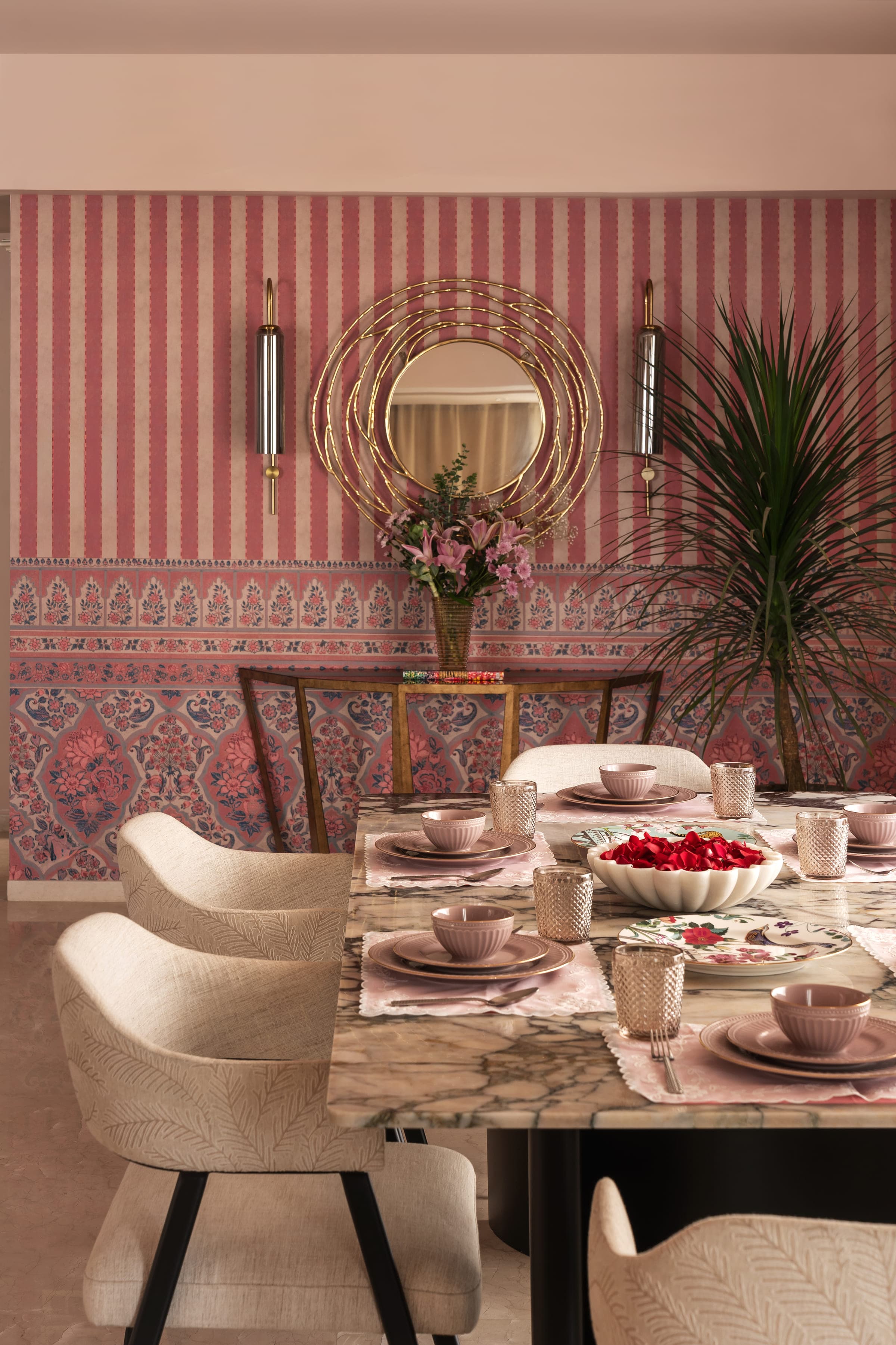 A dining table laid with pale crockery in front of a wall papered in a rose and green block print, a round mirror above the console.