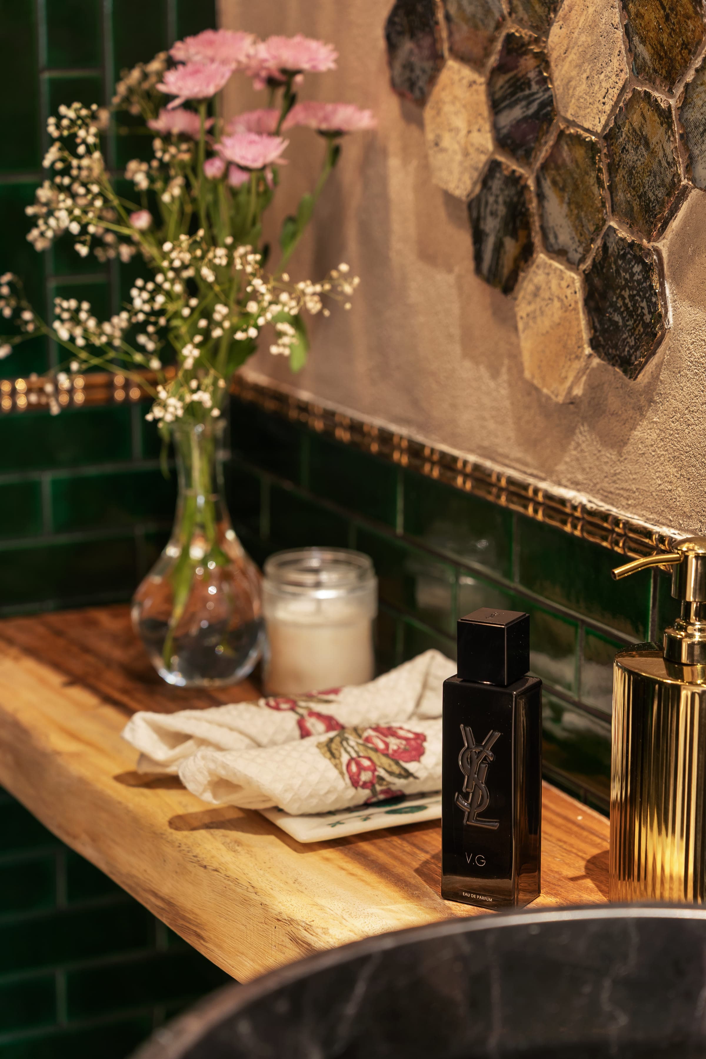 A detail of the green-tiled bathroom: a glass vase of gypsophila and a lit candle on a brass ledge.