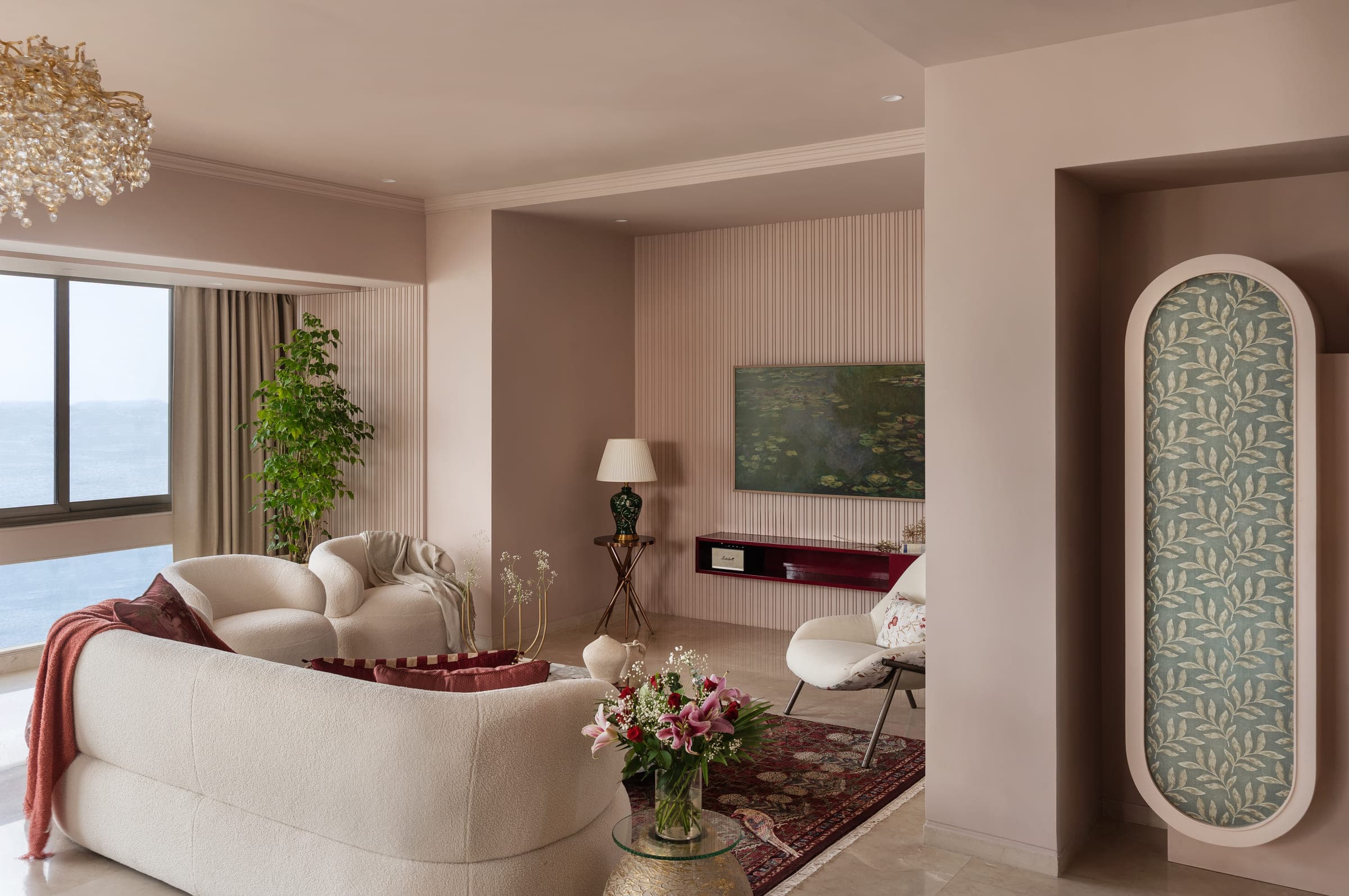 The living room of a sea-facing Bandra apartment: curved cream seating, a fluted blush wall, a burgundy lacquered console and full-height glazing onto the water.