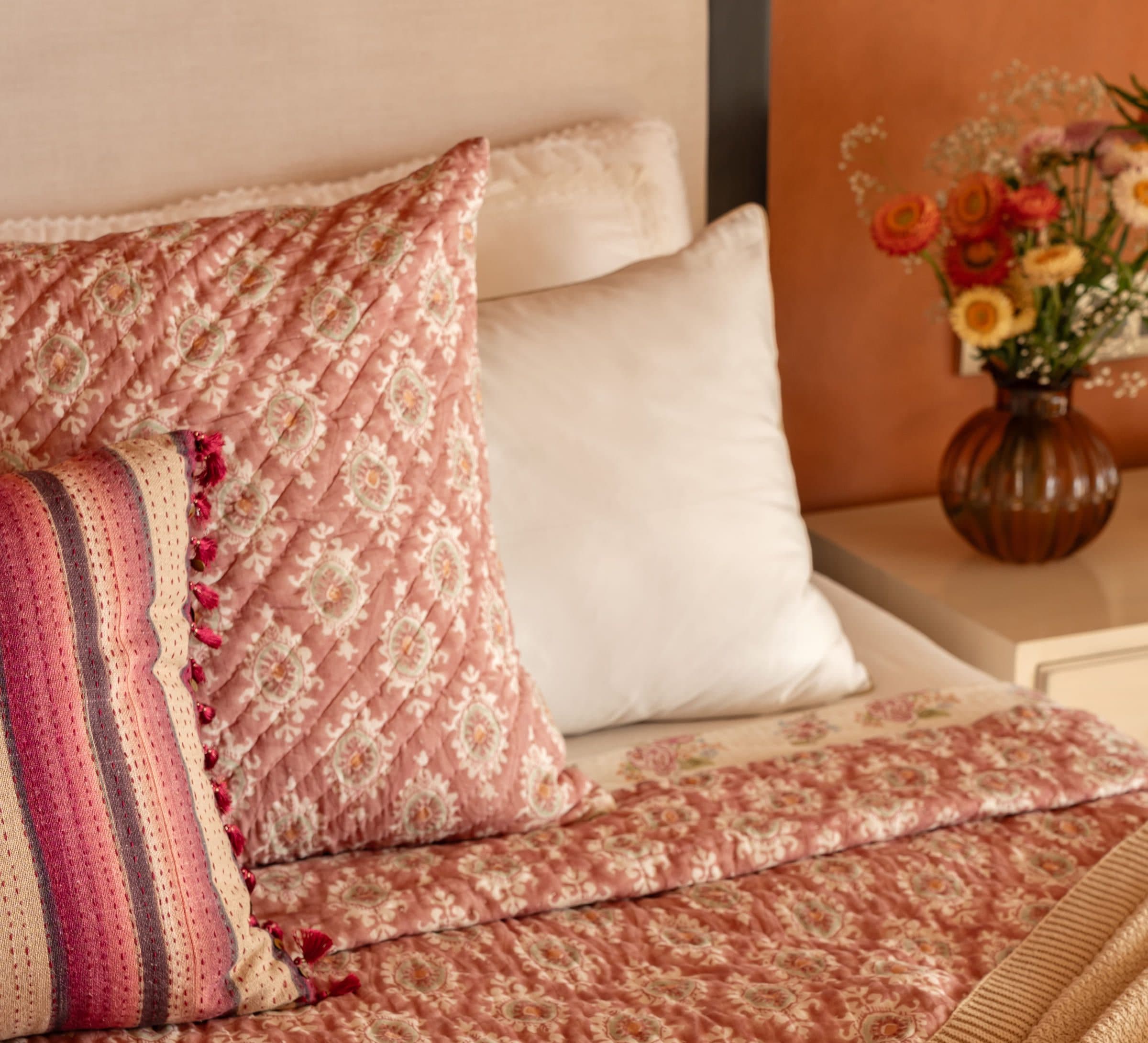 A close view of layered bedding: a block-printed quilt, a tasselled cushion and crisp white linen.