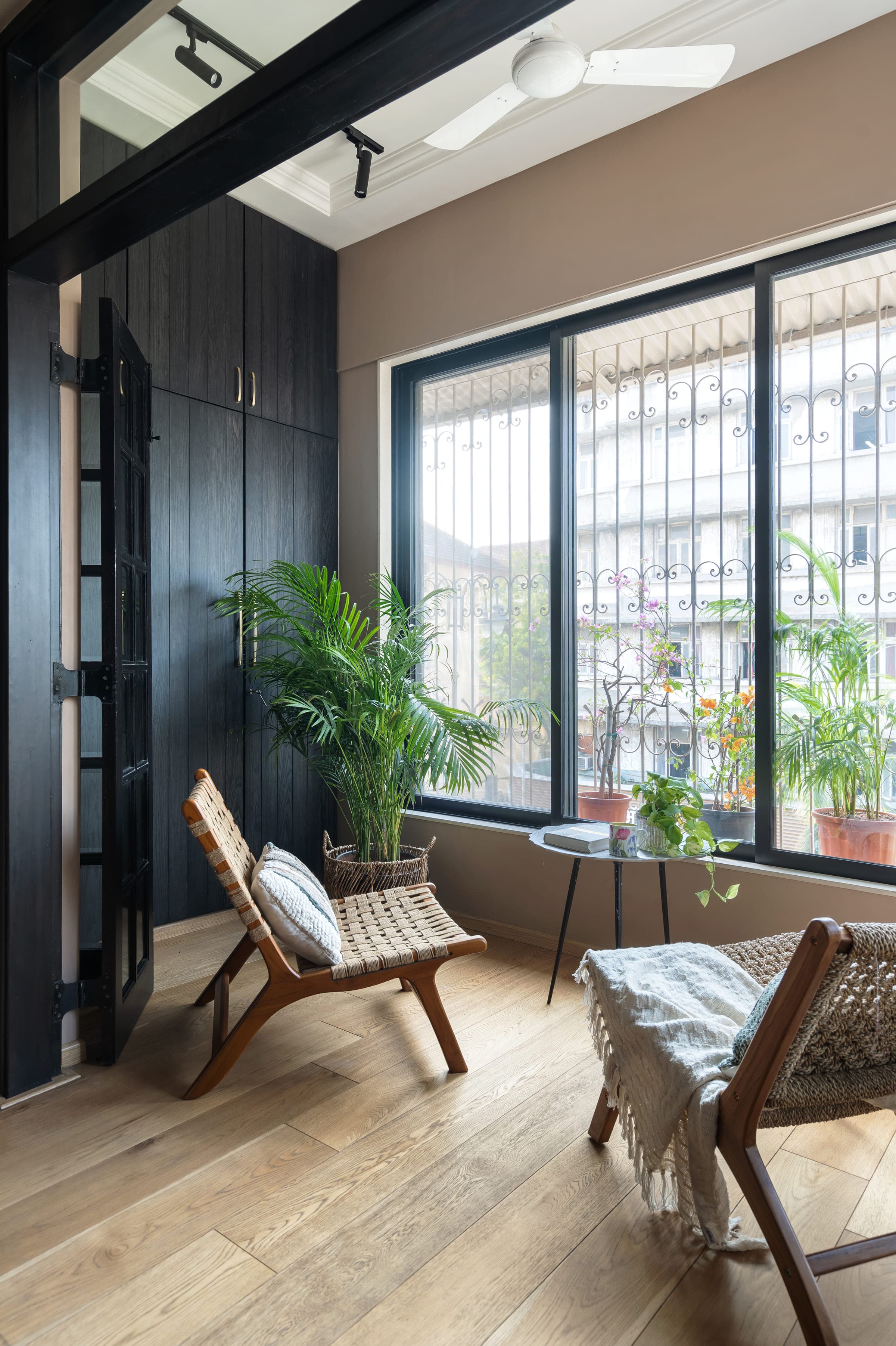 A balcony corner behind dark timber doors, potted plants along the sill and a cane chair beside them.
