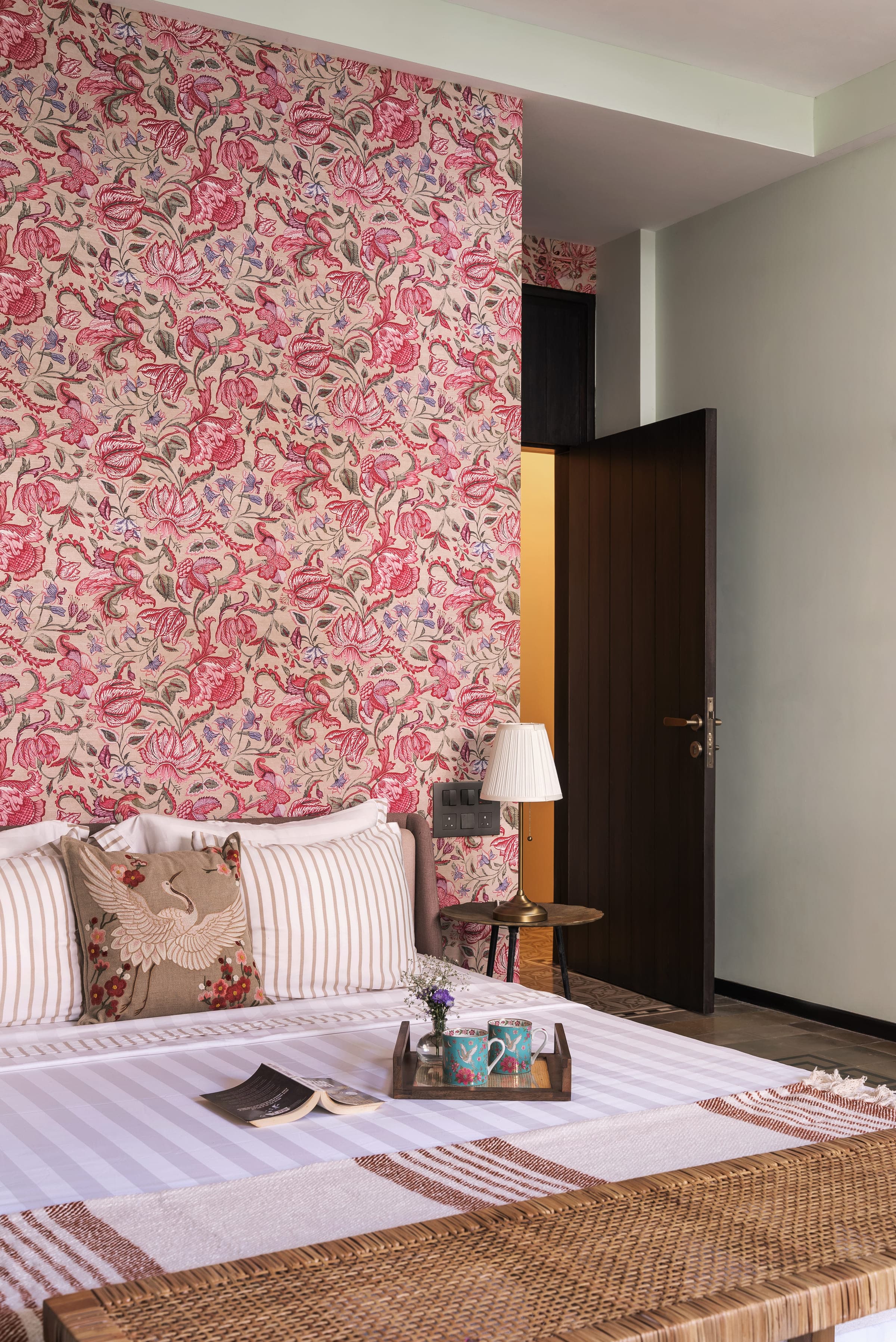 A bedroom with one wall papered in a large coral and rose botanical print, striped bedding, an embroidered crane cushion and a cane bench at the foot of the bed.
