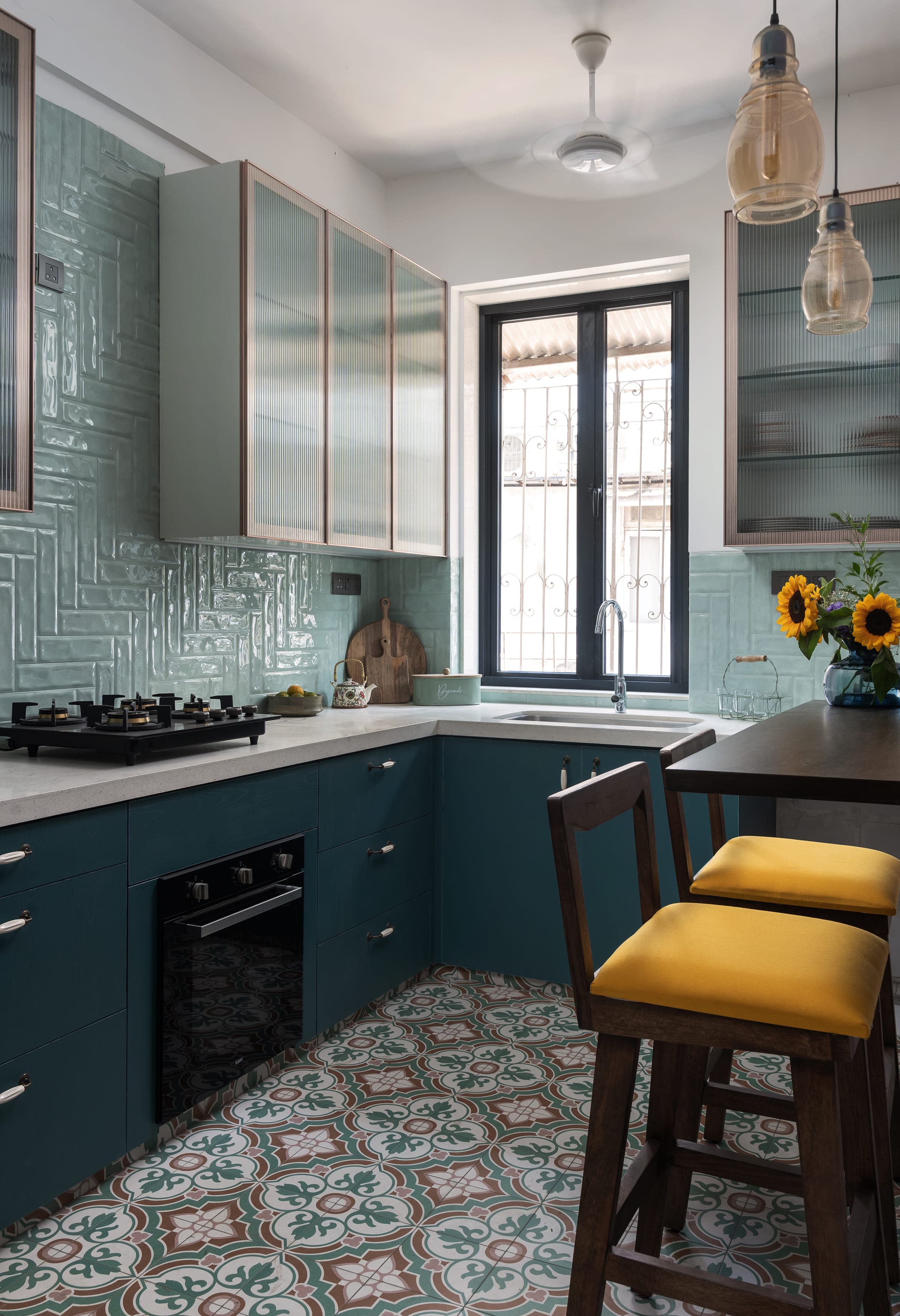 A kitchen in deep teal with glazed pale-blue subway tiling, white counters, a gas hob and sunflowers on the windowsill.
