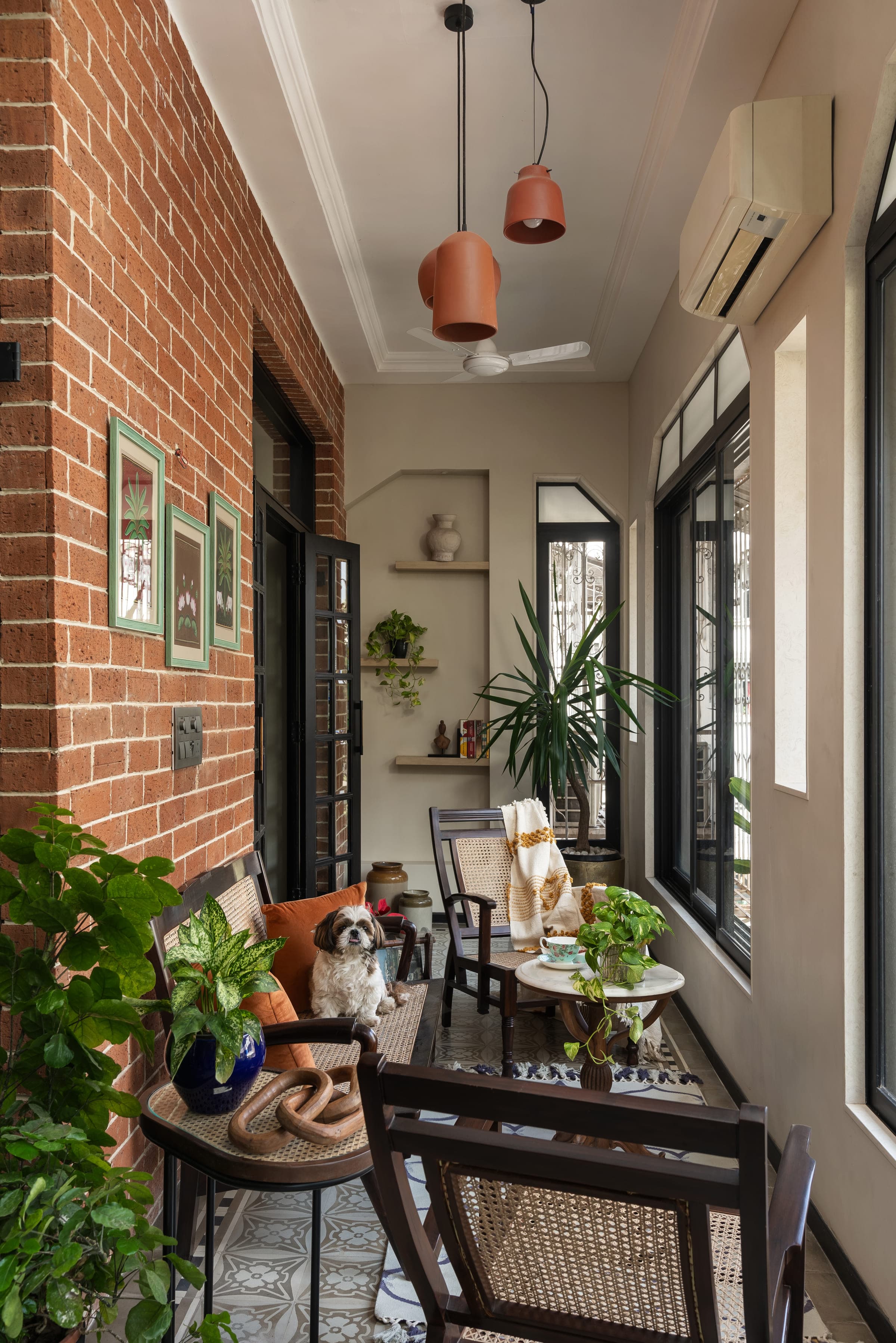A verandah lined in exposed brick with framed Pichwai paintings, terracotta pendant lights, cane and dark timber seating, potted plants and a patterned blue and white tile floor.