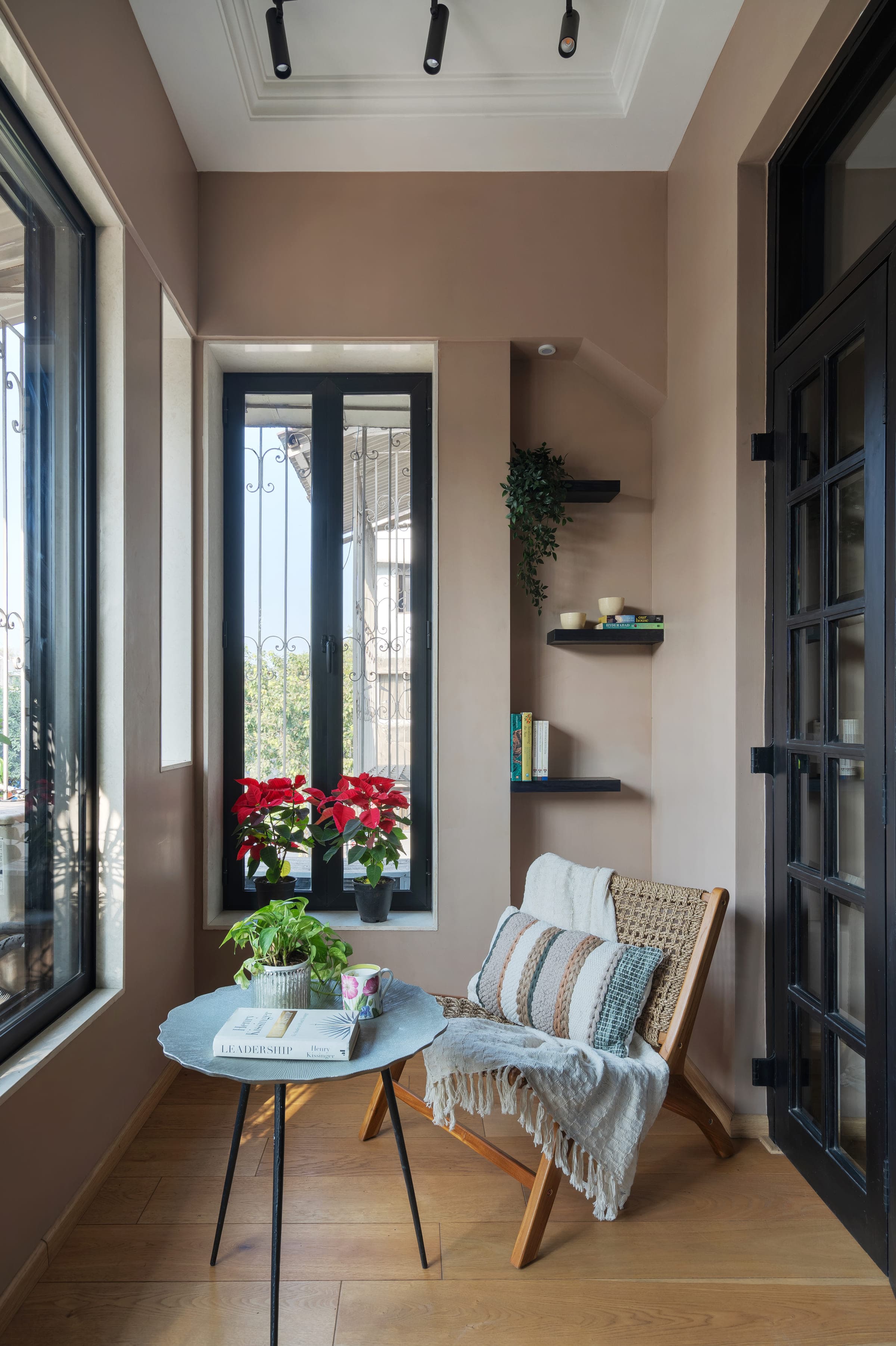 A reading corner by the window: a cane and timber chair with a throw over it, a pale blue table with books, poinsettias on the sill and black glazed doors alongside.