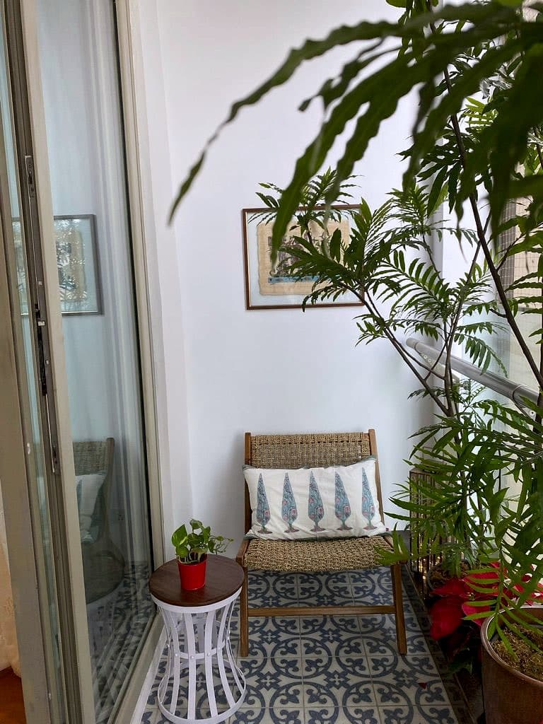 A cane chair on a patterned tile balcony floor, surrounded by ferns and palms, seen through the open bedroom door.