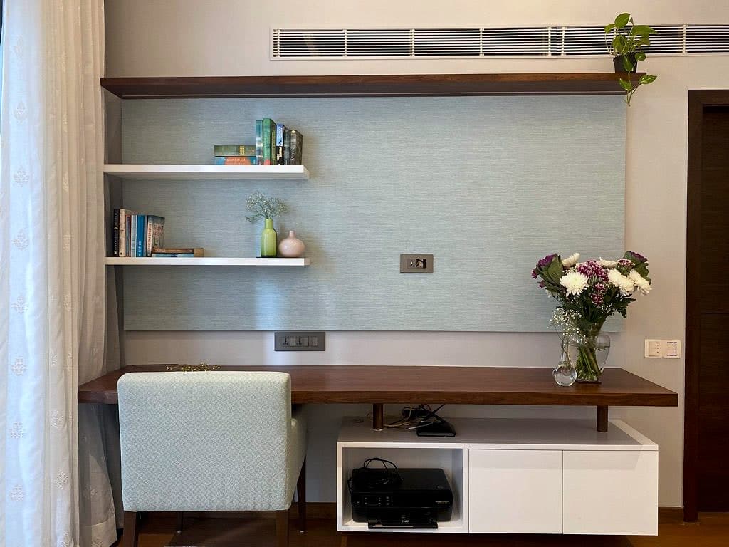 A teak desk shelf under two floating shelves of books, a pale upholstered stool tucked beneath and white cabinets alongside.