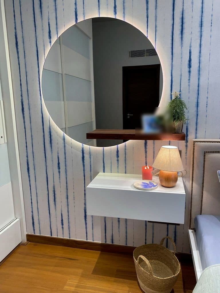 A round backlit mirror above a floating white console, a small lamp and framed photograph on it, striped wall behind.