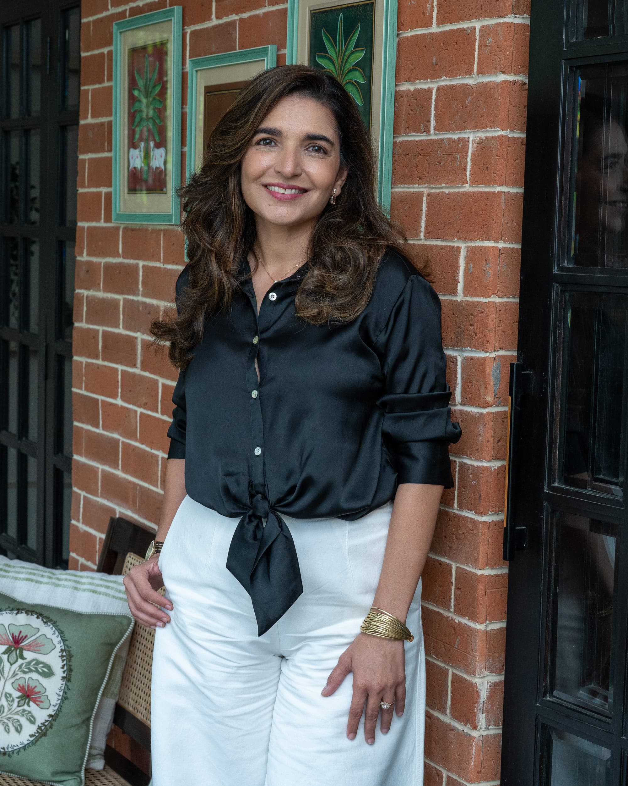 Azmin Kaka standing against a red brick wall hung with framed botanical tilework, beside a black-framed glazed door, in a black shirt and white trousers.